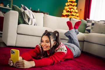 Obraz premium Young woman in festive sweater taking selfie on carpet near Christmas tree in cozy home during holiday season