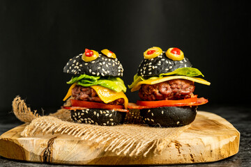 Close-up of two monster-style mini hamburgers with black buns and olive eyes on burlap over wooden board, top view food photography for Halloween or kids menu