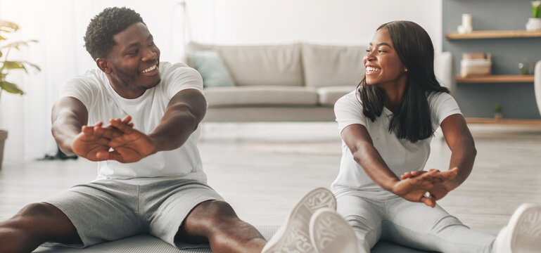 Happy young African American couple stretching sitting on floor yoga mats, making domestic workout, doing flexibility exercises at home. Fit black guy and his girlfriend training in living room