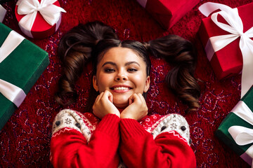 Joyful young woman in a festive sweater surrounded by colorful gift boxes, enjoying the cheerful...