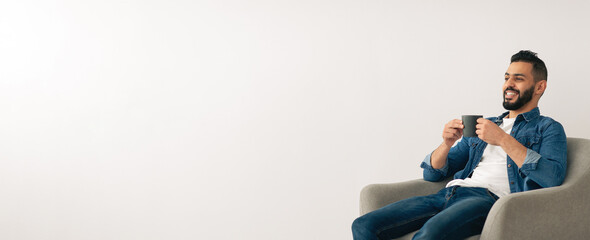 A man sits relaxed in a soft chair, sipping coffee with a smile. The bright room has a simple design, creating a calm and inviting atmosphere. He enjoys his moment of peace and joy.