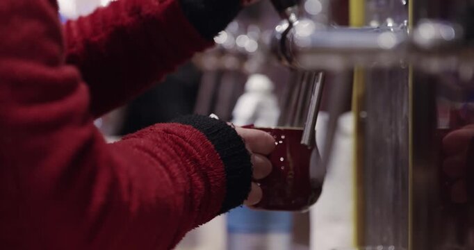 Person in a red jacket is pouring a hot wine Christmas drink into a cup at "Winemarkt" in Germany. The person is wearing a black glove on their left hand