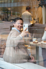 Smiling woman takes a photo with her smartphone through a cafe window, surrounded by coffee cups and pastries