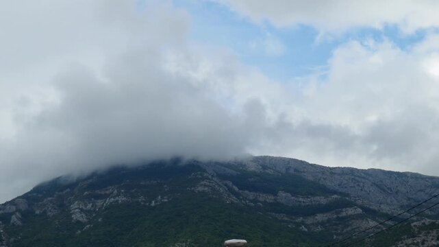 Clouds roll over the rugged mountain, creating a captivating view. The lush greenery contrasts with the grey rock faces under the vast sky. A tranquil moment in nature unfolds. Montenegro, Canj, Bar