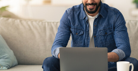 A man with a beard is sitting comfortably on a couch, engaged in his work on a laptop. He seems...