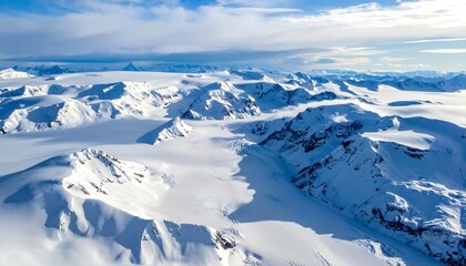 Aerial view reveals snow-covered mountain range under partly cloudy skies. The peaks and valleys are blanketed in white, with a hint of blue