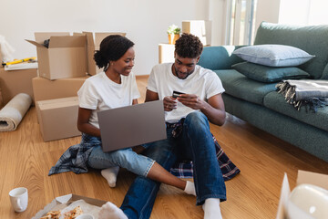 Smiling couple sitting on the floor of their new home, using a laptop and credit card to shop or...