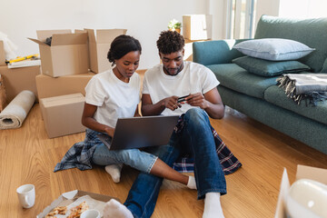 Smiling couple sitting on the floor of their new home, using a laptop and credit card to shop or...