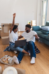 Joyful couple sitting among moving boxes, celebrating a successful online purchase or home setup while using a laptop during their move into a new home.