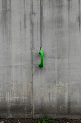 A vibrant, bright green telephone receiver hangs from its coiled black cord against a stark, weathered concrete wall, creating a powerful visual contrast. The rough, textured grey surface of the wall,