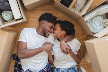 Happy African American couple lying on the floor surrounded by moving boxes, holding hands and smiling lovingly, celebrating new beginnings and the joy of moving into their first home together