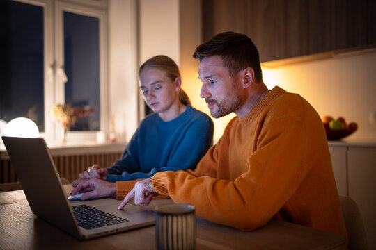 Exhausted young couple telecommuting, browsing and working on laptop in kitchen at night. Boyfriend and girlfriend electronic learning together, surfing internet, remote work together at night
