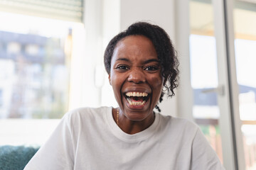 Portrait of a cheerful woman smiling brightly at home. Natural light, casual white shirt, and relaxed setting convey happiness, confidence, and positive energy. Having a video call. 