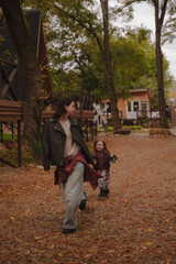 Fototapeta premium Mother and daughter run around the autumn yard near cozy A-shaped houses. The girls appear to be dressed in leather jackets, blue jeans and plaid shirts. Autumn atmosphere. Vertical photo.