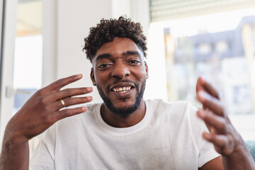 Smiling man having a videocall at home, talking and gesturing expressively while wearing a casual white t-shirt. Bright natural light and relaxed atmosphere convey friendliness and confidence