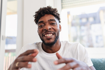 Smiling man having a videocall at home, talking and gesturing expressively while wearing a casual white t-shirt. Bright natural light and relaxed atmosphere convey friendliness and confidence