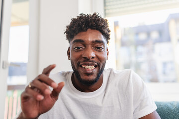 Smiling man having a videocall at home, talking and gesturing expressively while wearing a casual white t-shirt. Bright natural light and relaxed atmosphere convey friendliness and confidence
