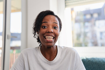 Portrait of a cheerful woman smiling brightly at home. Natural light, casual white shirt, and relaxed setting convey happiness, confidence, and positive energy. Having a video call. 
