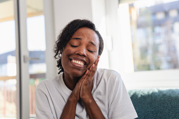 Young African American woman sitting indoors during a video call with a friend or family member, smiling warmly and reacting emotionally in a relaxed home environment