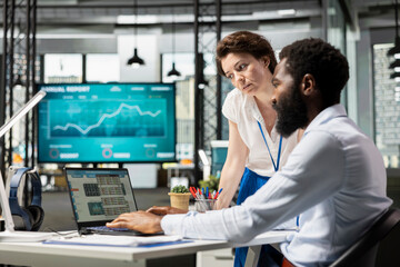 Female team leader and male staff discussing innovative strategies, evaluating data and insights in laptop. Diverse colleagues in office, working as team to improve outcomes and foster company growth.