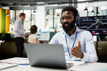 Business consultant wearing headphones engaging in video conference on laptop, explaining company strategies to remote clients. Black male manager in office, leading online meeting.