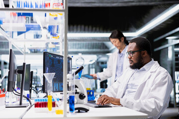 High tech lab technician at desk typing on PC keyboard, looking at 3D molecular model. African american man using bioengineering program on PC in medical lab displaying DNA data