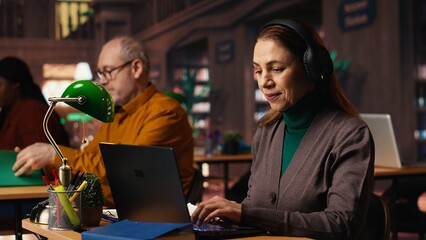 Mature student listening to school webinar on laptop at a library, takes notes and does research during online lecture. Studying remotely with an expert to obtain her diploma.