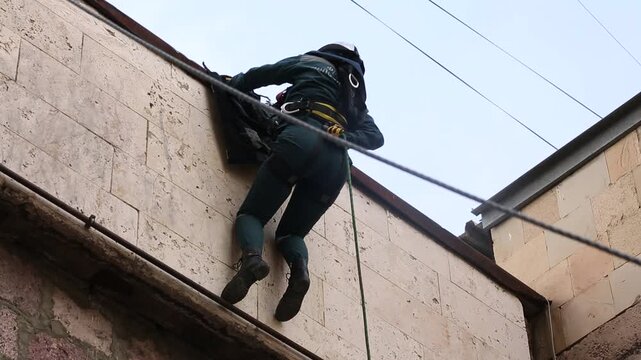 A professional industrial climber in safety harness and uniform rappels down the exterior brick wall of a building. Demonstrates high-rise rope access skills and worker safety training.