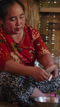 Asian woman peeling vegetables sitting on the floor cross-legged in a traditional kitchen with bamboo walls