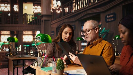 Mature college students completing class notes on papers and tablet, writing important information in a study area under green lamp. Colleagues pursuing academic qualifications.