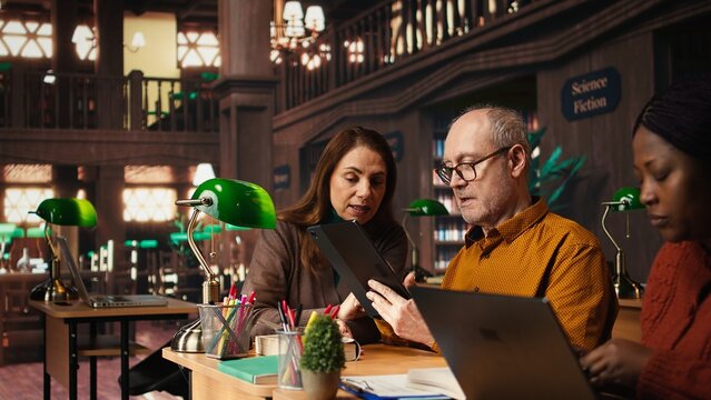 Mature college students completing class notes on papers and tablet, writing important information in a study area under green lamp. Colleagues pursuing academic qualifications.