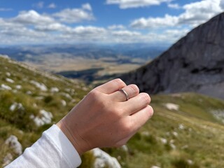 Landscape photo showing a sleek silver wedding band on a hand with bright natural scenery and...