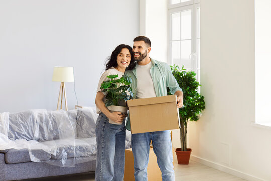 Happy couple moving, unpacking cardboard boxes in new home apartment. Young man and woman celebrate first day of relocation with smile, people holding potted plant and cartons, standing together