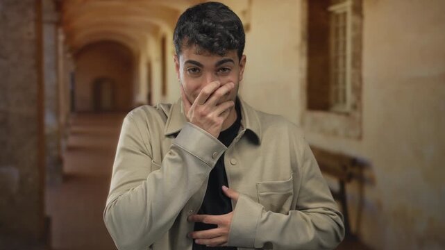 Man presses mouth then hides face in corridor where young hispanic actor shows surprise after laugh triggers expression familiar to every guy.