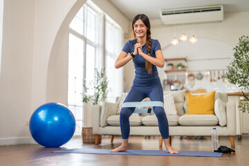 Fit smiling young woman doing exercise resistance band squats at home on yoga mat at home workout, healthy lifestyle, cardio fitness and strength training, bodyweight training and warm up, wellness