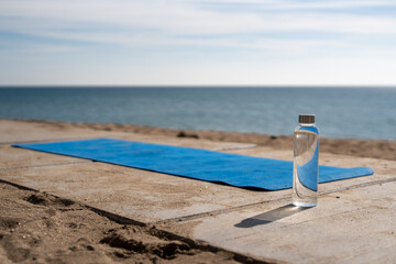 Transparent water bottle and blue yoga mat on sandy beach near sea, symbolizing wellness, mindfulness, hydration, and peaceful outdoor fitness in natural coastal environment