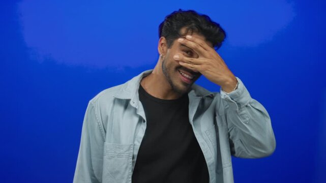 Young hispanic man covers face with hand and smiles through fingers in studio setting; bashfulness play.