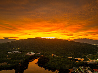A breathtaking aerial view of a vivid orange sunset casting dramatic light over forested hills and a reflective river, with urban elements nestled at the landscape’s edge beneath a glowing sky.