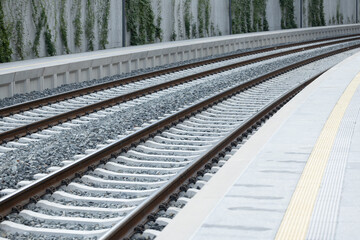 Railway tracks with gravel and concrete, curving gently along a modern station platform, surrounded by greenery and urban architecture, showcasing transportation infrastructure