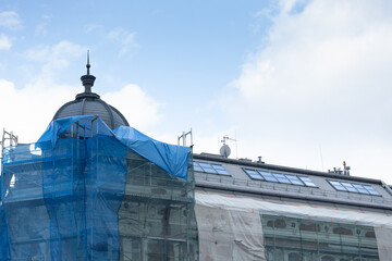 Building restoration project featuring scaffolding and protective blue netting, showcasing architectural details and a dome against a bright sky, highlighting urban development efforts