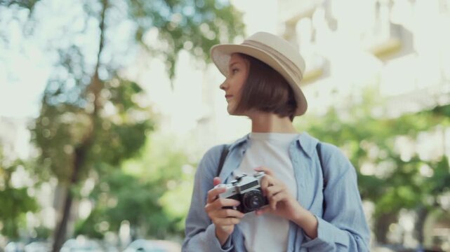 Young tourist girl in straw hat on summer city street playfully taking photo with retro camera during travel