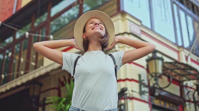 Happy young traveler girl in straw hat with backpack raising hands in relaxing gesture enjoying city street view near hotel on vacation