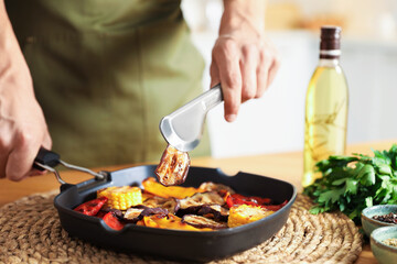 Man taking piece of tasty eggplant from pan with grilled vegetables at table indoors, closeup