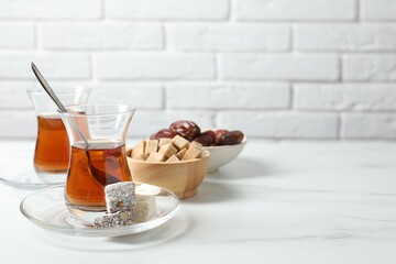 Tasty Turkish tea in glass cups, brown sugar and sweets on white marble table against brick wall, closeup. Space for text