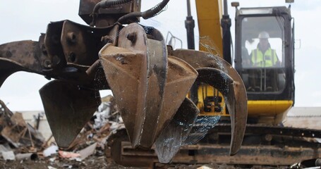 Hovering rusted mechanical grapple claw over scrap metal piles in salvage yard, with excavator cab