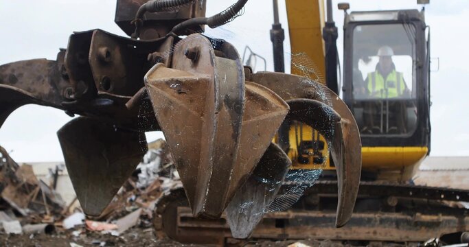 Hovering rusted mechanical grapple claw over scrap metal piles in salvage yard, with excavator cab