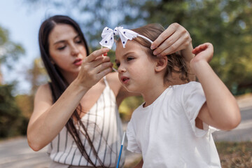 Mother adjusting a hair accessory for her young daughter during a sunny outing in a park setting