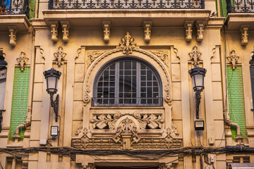 Facade of an ornate historic building in Alicante, Spain, featuring elegant wrought iron balconies, pastel green tiles, and decorative window moldings in modernist style.