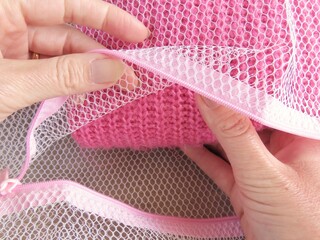Woman hands taking a pink knitted tricot out of a white laundry bag. Rustic wooden background. Organization concept.	