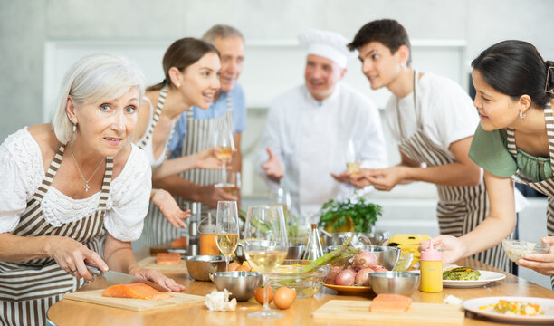 Pleased old woman learner of culinary classes processing piece of salmon standing around kitchen table together with others